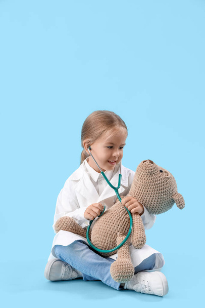 Χαριτωμένο μικρό γιατρό με στηθοσκόπιο και αρκουδάκι σε μπλε φόντο - Φωτογραφία, εικόνα