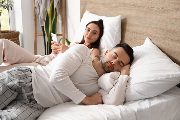 Asleep young man and his wife using mobile phone in bedroom - Photo, Image