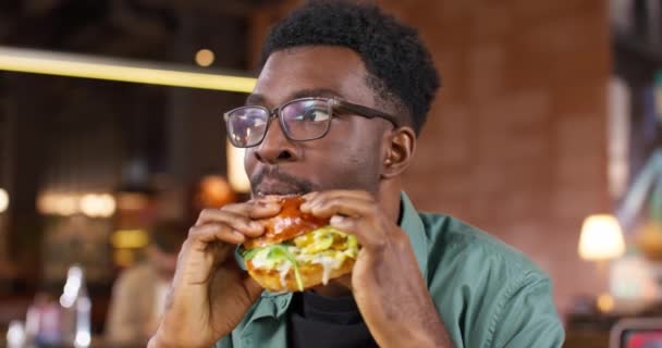Homem afro-americano faminto comendo hambúrguer saboroso no café ou restaurante. Jovem macho com óculos desfrutando de deliciosa comida. Sorrindo de alegria enquanto segurando em mãos fast food. Comida de rua. - Filmagem, Vídeo