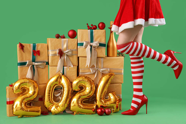 Femme sexy en bas de Noël debout près de la pile de cadeaux et figure 2024 faite de ballons de papier d'aluminium sur fond vert - Photo, image
