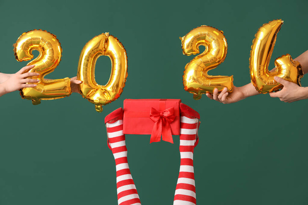 Χέρια με εικόνα 2024 κατασκευασμένα από μπαλόνια φύλλο, γυναικεία πόδια σε χριστουγεννιάτικες κάλτσες και κουτί δώρου σε πράσινο φόντο - Φωτογραφία, εικόνα