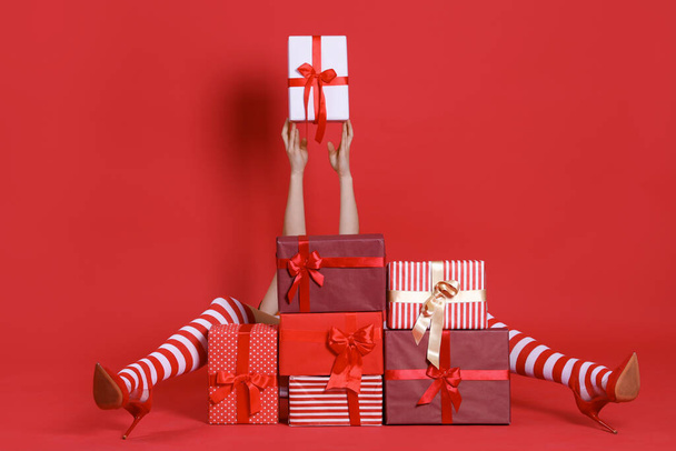 Frau in Weihnachtsstrümpfen sitzt hinter Stapel von Geschenken und hält Geschenkbox auf rotem Hintergrund - Foto, Bild
