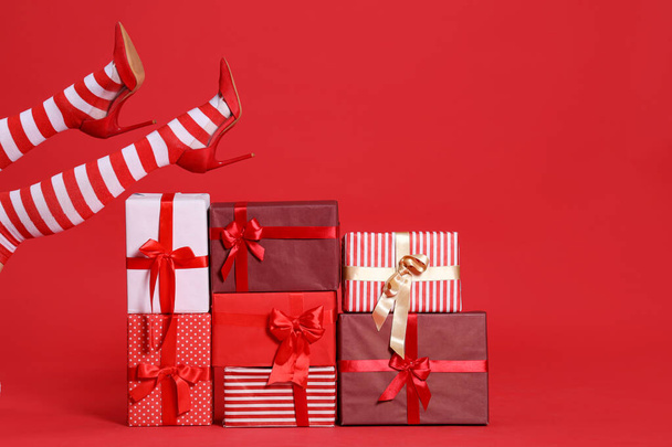 Female legs in Christmas stockings with stack of presents on red background - Photo, Image