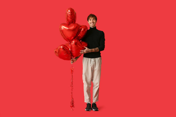 Joven guapo con globos de aire en forma de corazón sobre fondo rojo. Celebración de San Valentín - Foto, Imagen