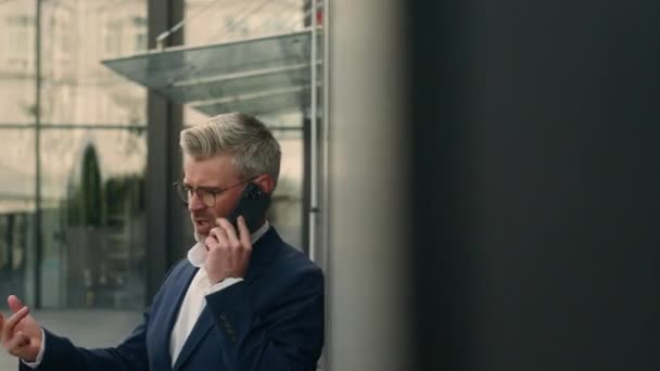 Angry 50s Caucasian worried man argue talking smartphone mad in city leaning wall of company office building stressed old businessman answer call mobile phone talk problem business conflict outdoors - Footage, Video
