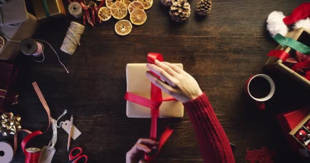 Boże Narodzenie, pakowanie prezentów i kłaniać się w ręce osoby z czerwoną wstążką na specjalne świętowanie. Powyżej drewniany stół, zbliżenie i pudełko na świąteczne wakacje, opakowanie niespodzianka prezent i kreatywna obsługa. - Materiał filmowy, wideo