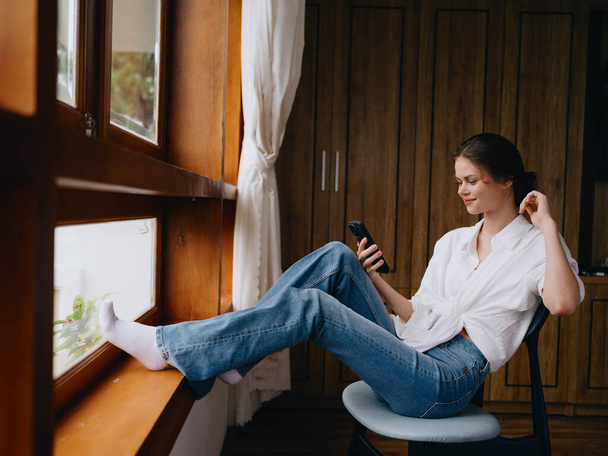 Jonge vrouw zit bij het raam met telefoon in de hand glimlach op zoek online, lente stemming, huis eenvoudige kleren, huis gezellige sfeer, esthetische levensstijl. Hoge kwaliteit foto - Foto, afbeelding