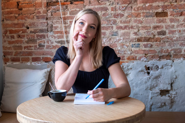 This image portrays a young woman with blonde hair sitting at a wooden table in a cafe. She appears contemplative, with one hand resting on her chin and the other holding a pen to paper. A cup of - Photo, Image