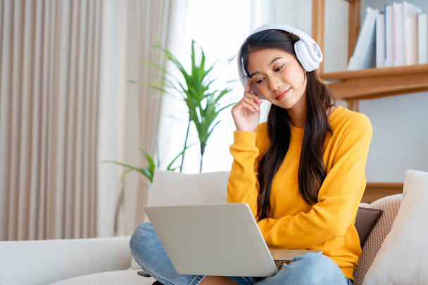 Nuori suloinen aasialainen nainen hymy yllään kuulokkeet työskentelevät olohuoneessa kotona. Onnellinen upea nainen tietokoneella kannettavan ja istuu sohvalla kotona, Videopuhelu ystävän kanssa - Valokuva, kuva