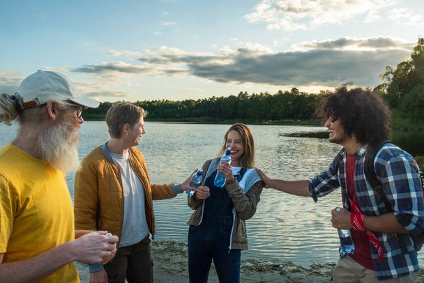 The photo depicts a cheerful moment among a group of friends by the lake, with an elderly Caucasian man, a young Caucasian man, an East Asian woman, and a Middle-Eastern man toasting with water - Photo, Image