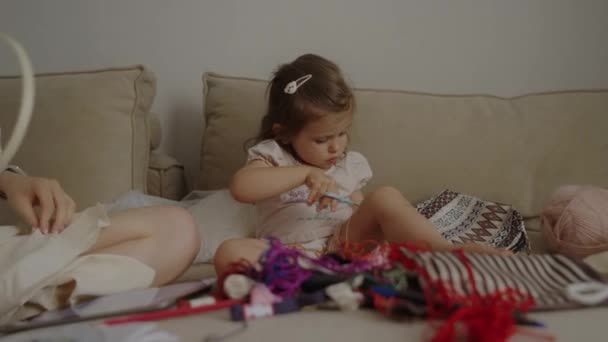 Konzentriert näht das süße kleine Mädchen nach Hause und sitzt neben ihrer Mutter auf dem Sofa. Kinder Vielfalt Glückserziehung Konzept. - Filmmaterial, Video