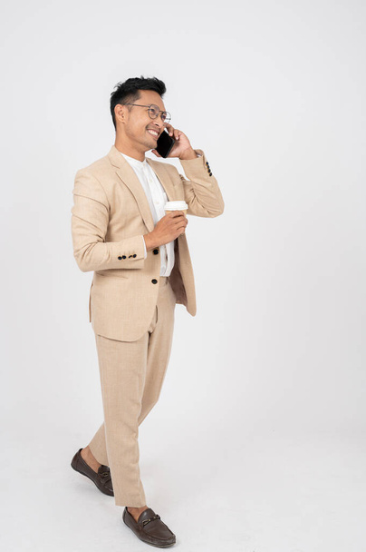 Un hombre de negocios asiático inteligente y positivo en un traje formal está hablando por teléfono y caminando con una taza de café en la mano, sobre un fondo de estudio blanco aislado. - Foto, imagen