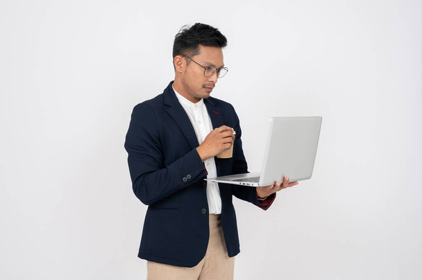 zestresowany, poważny azjatycki biznesmen w formalnym garniturze pracuje, stojąc na białym tle z komputerem na laptopie. - Zdjęcie, obraz