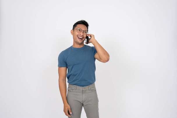 Radosny, szczęśliwy Azjata w luźnych ciuchach cieszy się rozmawiając z kimś przez telefon, stojąc na odizolowanym białym tle studia. technologia bezprzewodowa, rozmowy telefoniczne, komunikacja - Zdjęcie, obraz