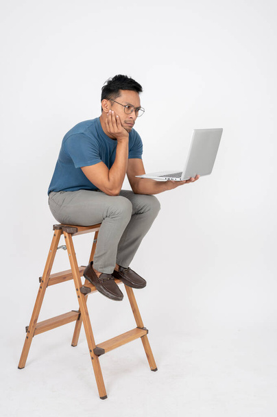 Sıkılmış Asyalı bir adam dizüstü bilgisayarında bir e-posta beklerken izole edilmiş beyaz bir stüdyo arka planında ahşap bir merdivende oturuyor. İnsanlar ve teknoloji kavramları - Fotoğraf, Görsel