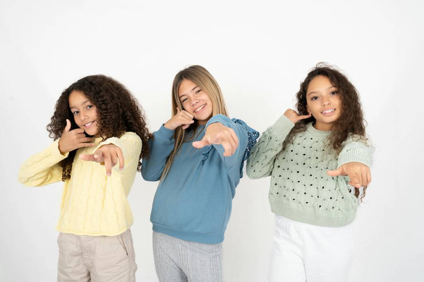Three young beautiful multiracial kid girls smiling cheerfully and pointing to camera while making a call you later gesture, talking on phone - Photo, Image