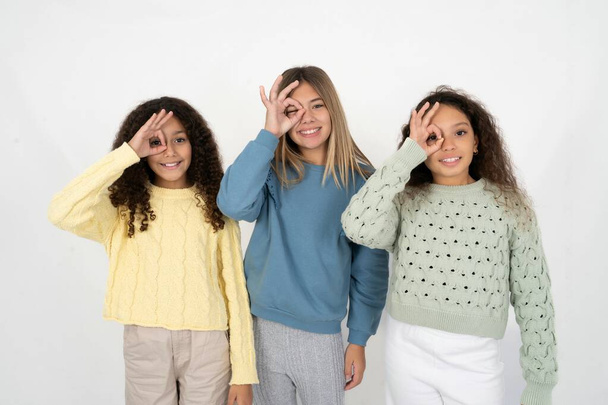 Tres chicas jóvenes hermosas multirraciales haciendo buen gesto con la mano sonriendo, ojos mirando a través de los dedos con la cara feliz. - Foto, imagen