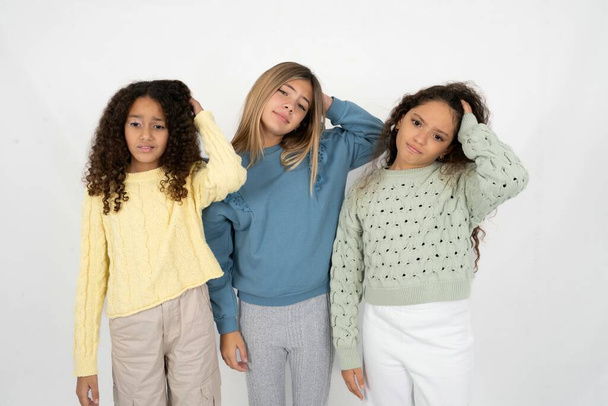 Three young beautiful multiracial kid girls being confused and wonders about something. Holding hand on head, uncertain with doubt. Pensive concept. - Photo, Image