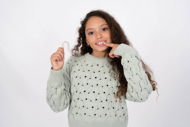 beautiful kid girl wearing sweater holding an invisible aligner and pointing perfect straight teeth. Dental healthcare and confidence concept. - Foto, Imagen
