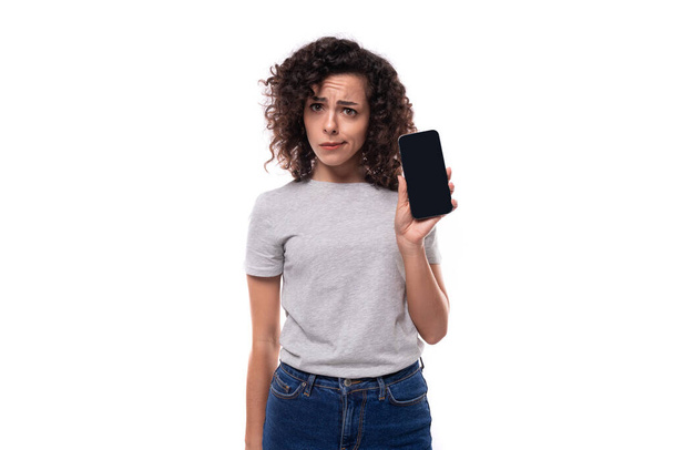 ładny młody kręcone brunetka kobieta w szary t-shirt pokazując smartfon ekran z makieta izolowane na białym tle z przestrzeń kopiowania. - Zdjęcie, obraz