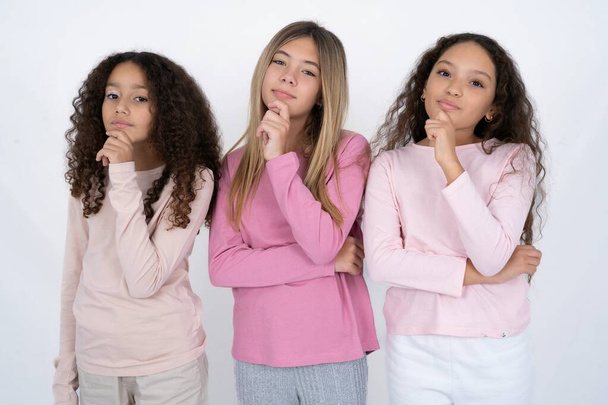 Τρία νεαρά όμορφα πολυφυλετικά κοριτσάκια που κοιτούν με αυτοπεποίθηση την κάμερα χαμογελώντας με σταυρωμένα χέρια και υψωμένα χέρια στο πηγούνι. Θετική σκέψη. - Φωτογραφία, εικόνα