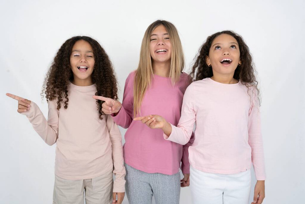 Three young beautiful multiracial kid girls laughs happily points away on blank space demonstrates shopping discount offer, excited by good news or unexpected sale. - Photo, Image