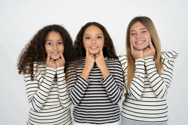 Ευτυχισμένα Τρία νεαρά όμορφα πολυφυλετικά κοριτσάκια με χαμόγελο στα δάχτυλα, κρατά δείκτη κοντά στο στόμα, δάχτυλα δείχνοντας και αναγκάζοντας το χαρούμενο χαμόγελο - Φωτογραφία, εικόνα