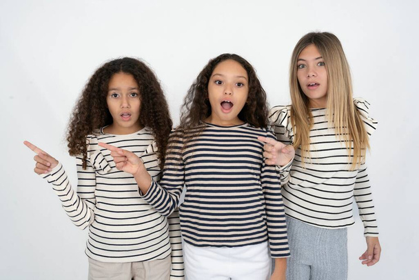 Emotive Τρία νεαρά όμορφα πολυφυλετικά κορίτσια παιδί κρατά το σαγόνι έπεσε από το σοκ δείχνει καταπληκτικό promo σημεία δεξιά στο κενό χώρο δείχνει μεγάλη πώληση αγορών. Διαφήμιση - Φωτογραφία, εικόνα