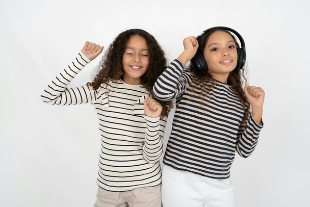 Carefree two young beautiful multiracial kid girls with toothy smile raises arms dances carefree moves with rhythm of music listens music from playlist via headphones - Фото, изображение