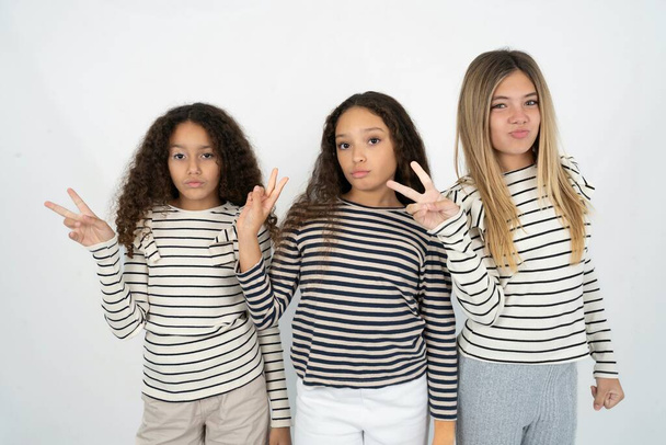 Three young beautiful multiracial kid girls makes peace gesture keeps lips folded shows v sign. Body language concept - Photo, Image