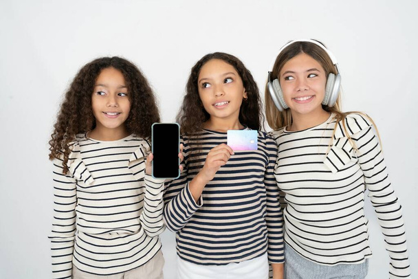 Drie jonge mooie multiraciale kid meisjes met bankkaart modern apparaat op zoek lege ruimte - Foto, afbeelding