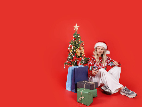 Hold karty kredytowej i smartfona, portret całego ciała siedzi Santa kobieta posiadają kartę kredytową i smartfona. Ozdobiona choinka, zapakowane pudełka, torby na zakupy. Płatność online, zakup. - Zdjęcie, obraz