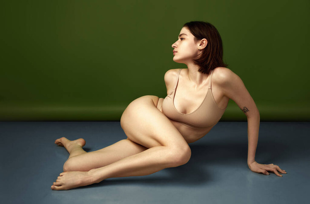 La bellezza della semplicità è catturata in questa immagine di giovane donna in lingerie color carne, posando con grazia sul pavimento dello studio sullo sfondo di oliva. Concetto di bellezza naturale, femminilità, benessere. - Foto, immagini