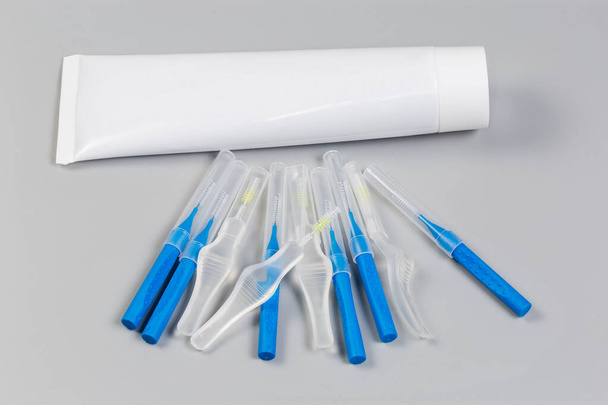 Diferentes cepillos interdentales desechables se encuentran en una superficie gris contra el tubo blanco de pasta de dientes - Foto, Imagen