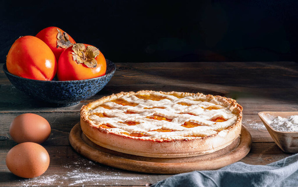 Italský domácí persimmon koláč nebo crostata s moukou, vejci a ovocem na dřevěné řezné desce a stole, zblízka, chiaroscuro styl, komfort potravin - Fotografie, Obrázek