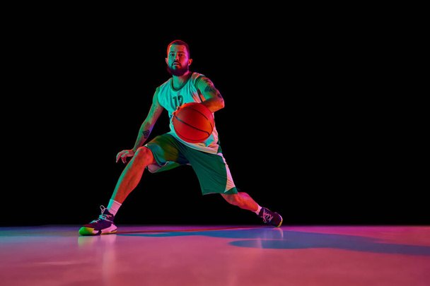 Full-length portrait of basketball player training dribbling technique and making powerful slam dunk against black background in neon. Concept of sport, hobby, active lifestyle. Copy space, ad - Photo, Image