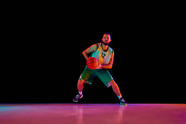 Retrato de comprimento total do homem atlético, jogador de basquete profissional ativamente treinando antes do jogo contra fundo preto em luz de néon. Conceito de esporte, hobby, estilo de vida ativo. Espaço de cópia, anúncio - Foto, Imagem