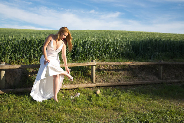 Νεαρή γυναίκα, όμορφη και ξανθιά με λευκό φόρεμα, κάθεται σε ξύλινο κιγκλίδωμα βάζοντας τα παπούτσια της, με ένα λιβάδι στο βάθος. Concept ομορφιά, μόδα, νύφες, παπούτσια, πόδια, αξεσουάρ. - Φωτογραφία, εικόνα