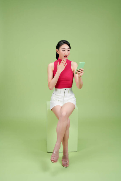 feliz sorprendida joven sentada usando su móvil aislado sobre fondo verde - Foto, imagen
