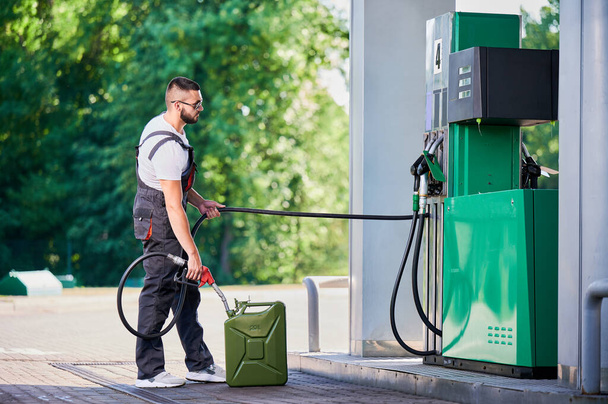 Professional worker refueling canister with fuel. Skilled man filling cistern with gasoline for customer. Man in ovealls with pump nozzle refueling canister next to gasoline column. - Photo, Image