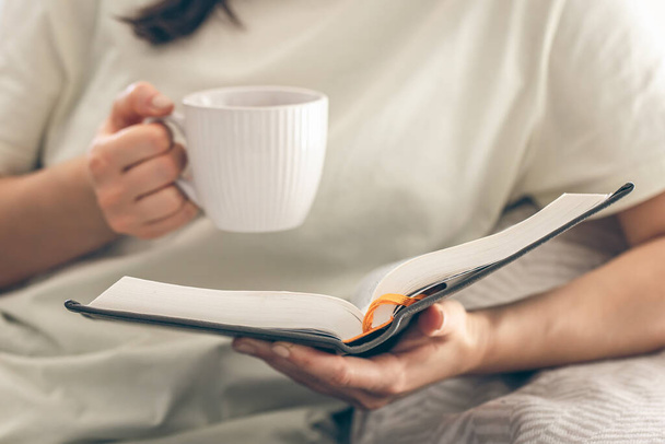 Jovem mulher lendo um livro em casa, relaxando no sofá. Close-up, livro e xícara em mãos femininas. - Foto, Imagem