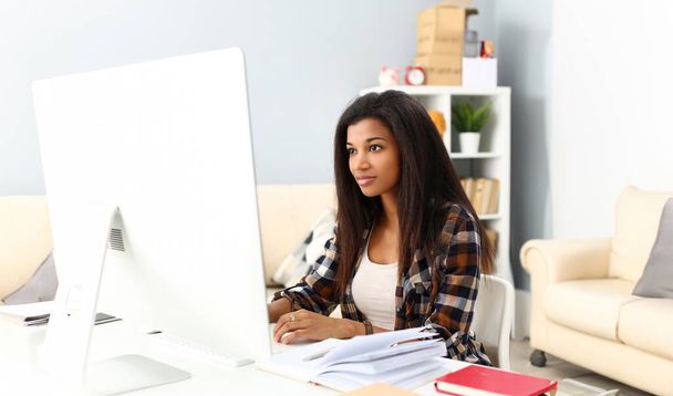 Femme souriante noire assise sur le lieu de travail travaillant avec ordinateur de bureau étudiant ou faisant des devoirs scolaires concept
 - Photo, image