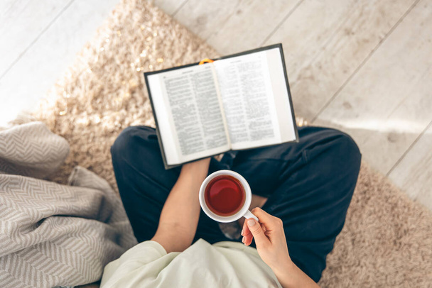 Vista dall'alto, donna che tiene una tazza di tè mentre legge un libro sul pavimento di un accogliente soggiorno. - Foto, immagini