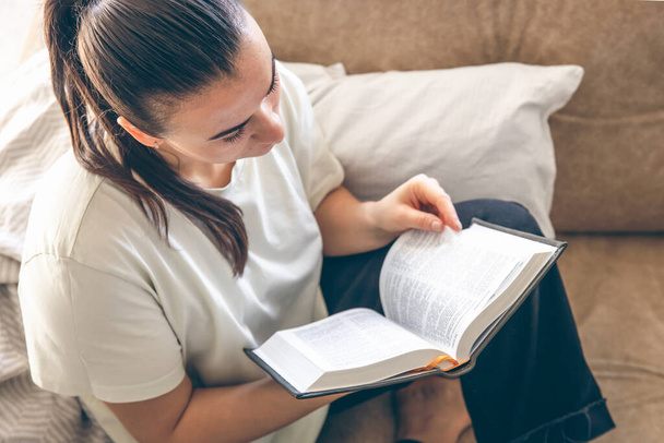 Young Caucasian woman reading a book at home. A woman is reading a book on the sofa. Hobbies, home leisure and a good reading habit. - Photo, Image