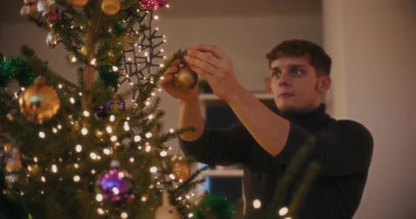 Młody mężczyzna wiąże bombkę podczas dekoracji podświetlanej choinki w domu - Materiał filmowy, wideo
