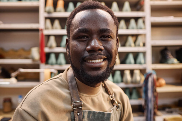 Fröhlicher junger afroamerikanischer Schuhmacher oder Lehrling blickt in die Kamera, während er in einer geräumigen Werkstatt mit Vorräten steht - Foto, Bild