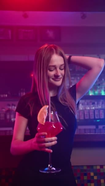Pretty Young Happy Girl Smiles and Dances in Nightclub with Red Cocktail in her Hands (en inglés). Antecedentes hay Bar Counter. Vídeo vertical, cámara lenta - Metraje, vídeo