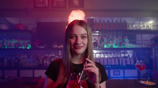 Ritratto di un'attraente ragazza sorridente che beve un cocktail rosso in un bar, nell'illuminazione al neon. Fiamme e fuoco sullo sfondo. Concetto di Relax e Intrattenimento di Notte. Rallentatore. - Filmati, video