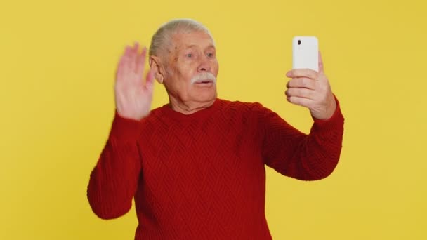 Happy senior old man blogger influencer taking portrait selfie on smartphone, comunicating record video call online with social media subscribers. Abuelo anciano aislado sobre fondo amarillo - Imágenes, Vídeo