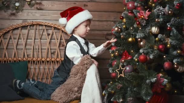 Leuke jongen in de kerstman hoed spelen met kerst ornamenten kerstballen op een kerstboom te wachten voor kerst te komen - Video